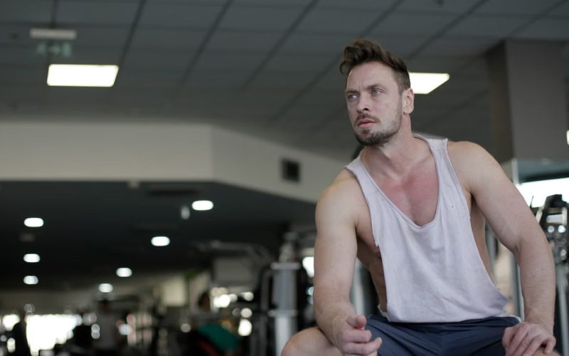 Muscular man with facial hair sitting and looking away in a gym, captured in selective focus.